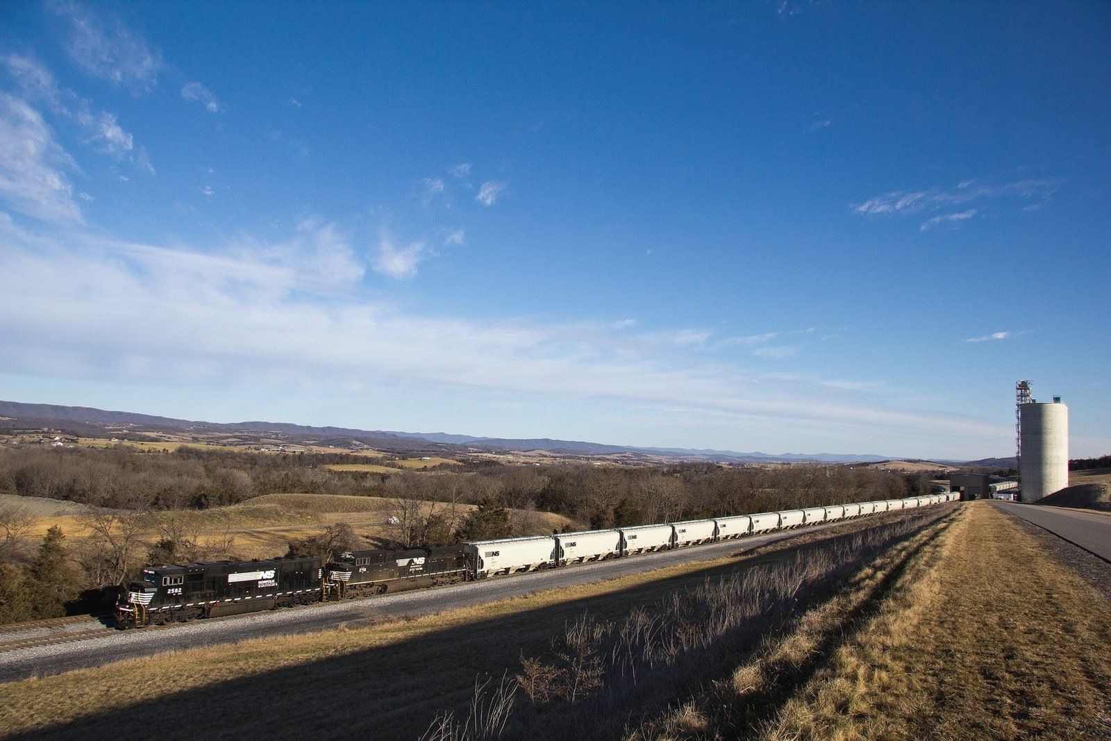 A long train of covered hopper cars running across open country toward a concrete elevator