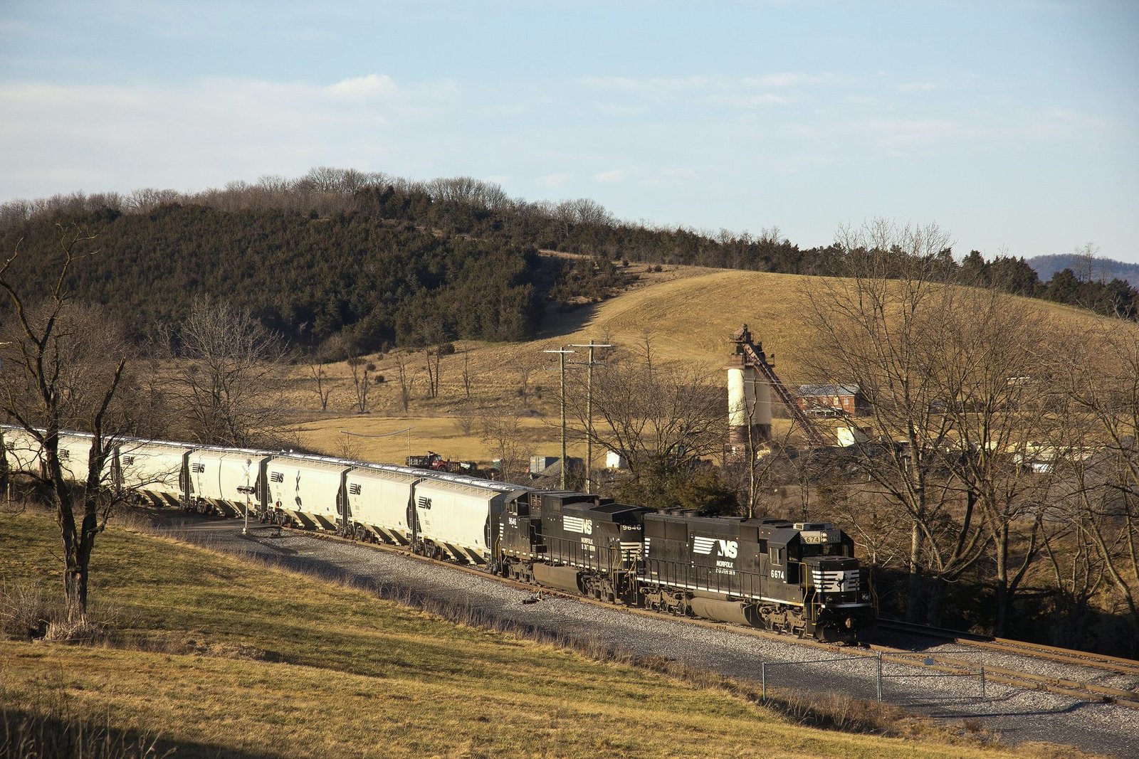 Locomotives pulling a long string of white hopper cars past a grain elevator in brown late-autumn country
